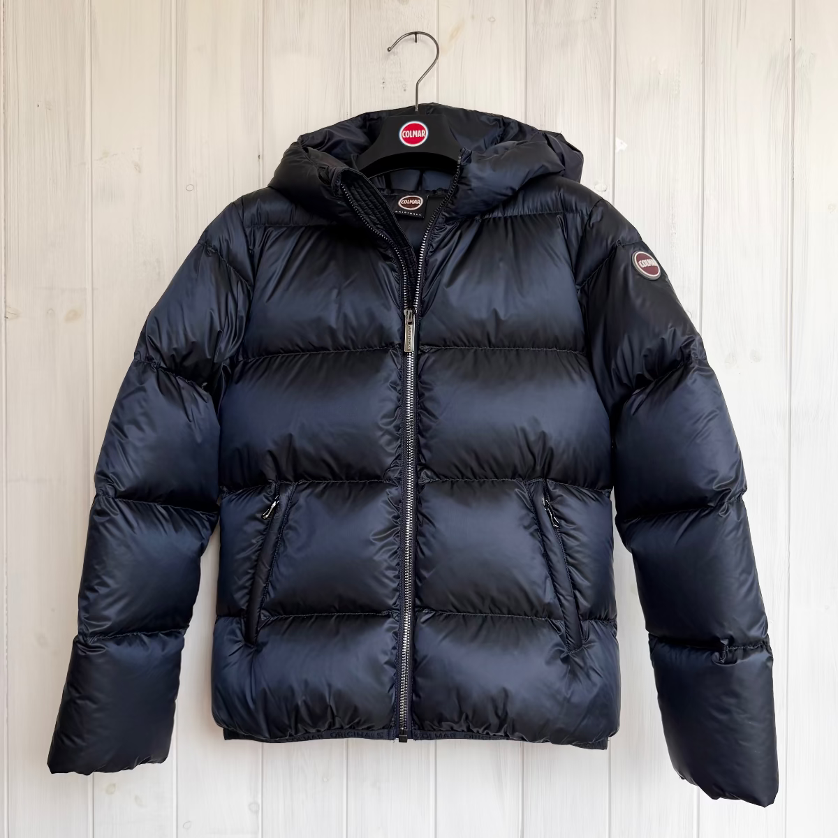 Navy puffer jacket from colmar on a hanger against a light wooden panel background