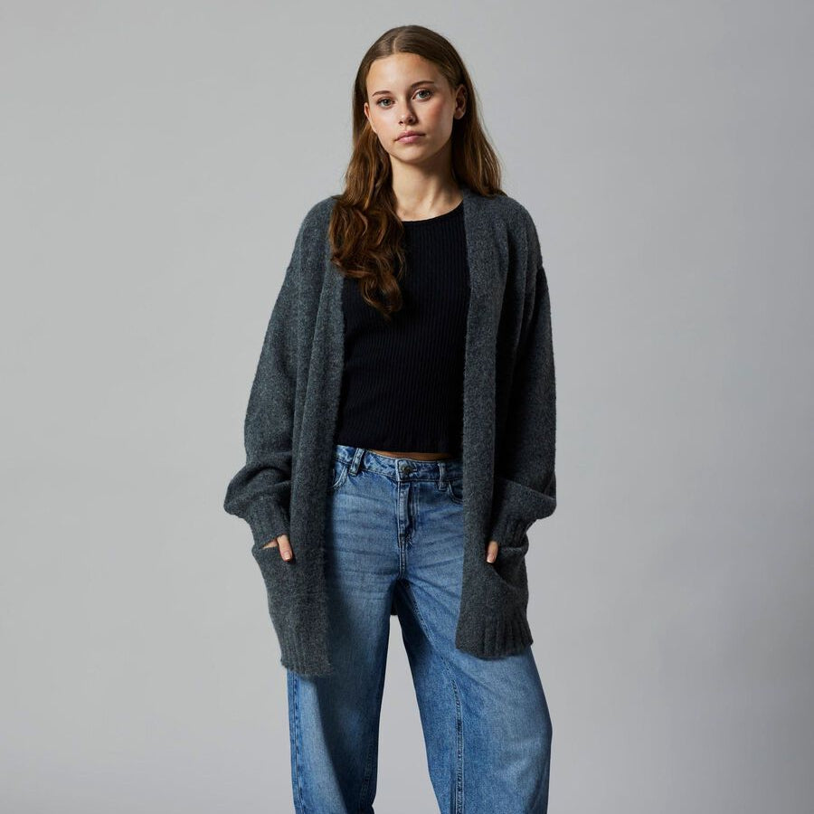 Woman wearing a dark gray cardigan, black top, blue jeans, and brown shoes on a gray background