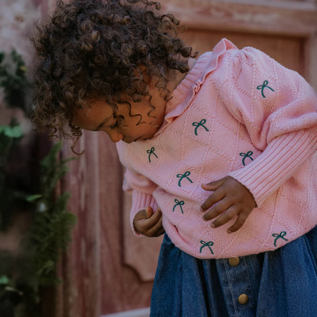 Child wearing a pink sweater with green ribbons and blue jeans, standing in front of a wooden door.
