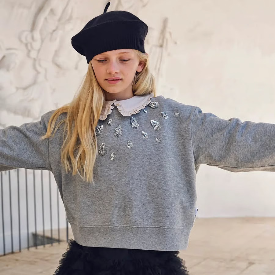 A young girl wearing a gray crewneck sweatshirt with rhinestone embellishments, a black tulle skirt, and a black beret, posing with her arms outstretched against a light-colored wall.