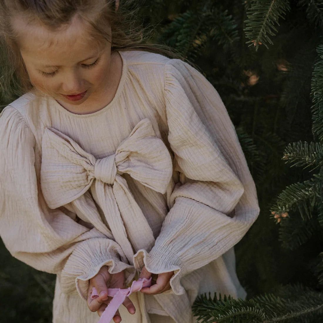 Young girl in a beige blouse with a large bow holding a pink ribbon, standing in front of a Christmas tree.