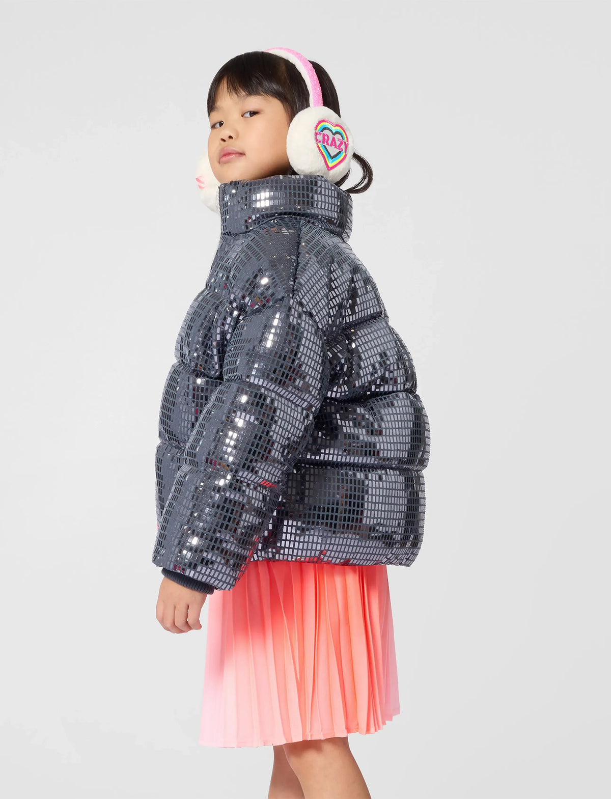 A child model wearing a shiny silver sequined puffer jacket, a pink pleated skirt, and white earmuffs with a pink band, photographed against a light gray studio background.