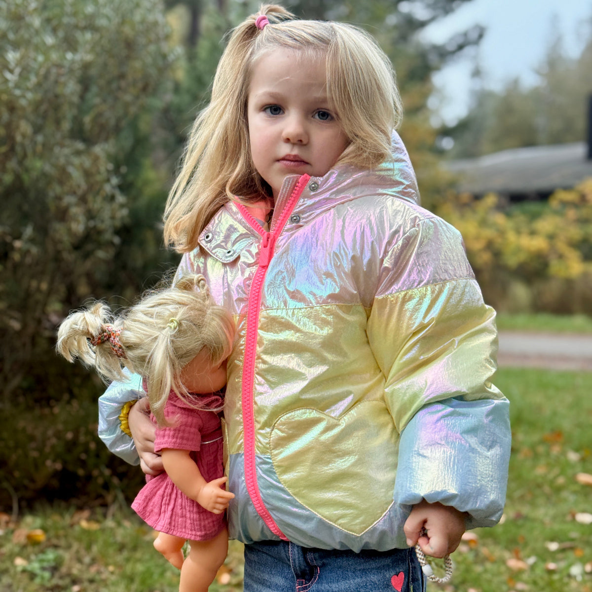 Young girl in rainbow puffer jacket holding a doll outdoors with a blurred background