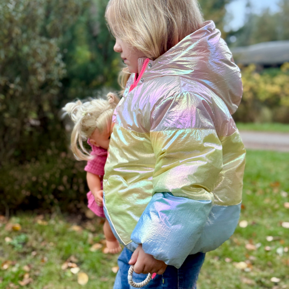 Young girl holding a doll wearing a colorful iridescent puffer jacket outdoors.