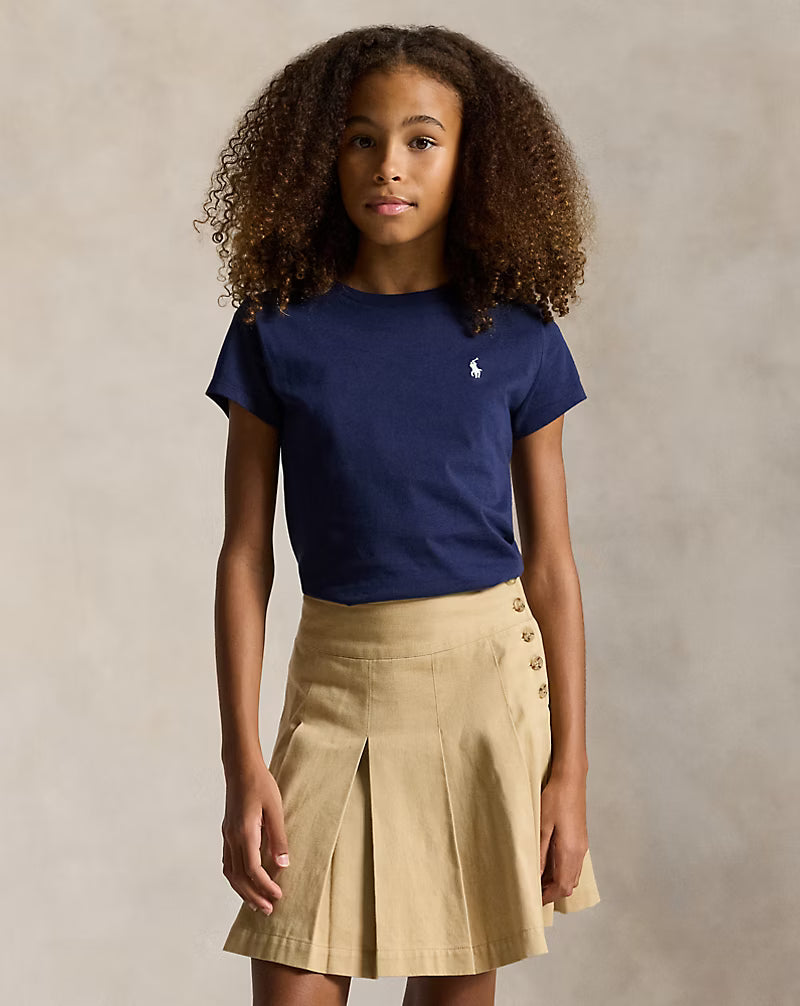 A young girl with curly brown hair wearing a navy blue short-sleeve t-shirt with a small white embroidered polo player logo on the chest, paired with a beige pleated skirt with button details, standing against a neutral textured beige background.