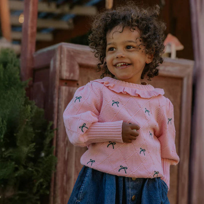 Child wearing a pink sweater with green bows and blue jeans standing outdoors.