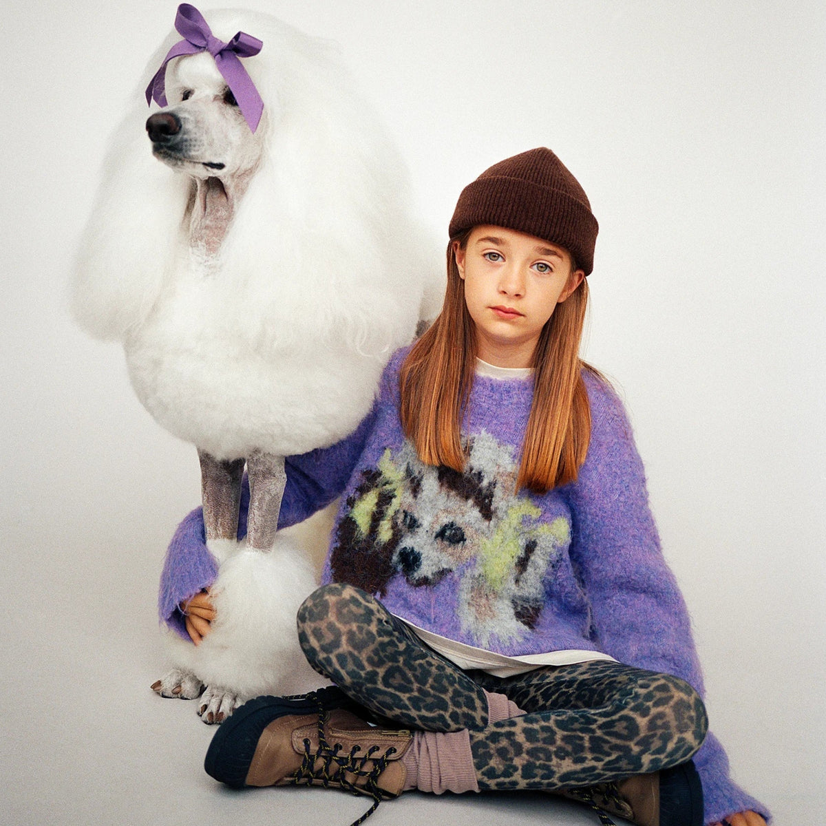 A young girl with long light brown hair sits cross-legged on a light gray studio floor next to a large white poodle. The girl wears a brown knit beanie, a fuzzy lavender sweater with a dog graphic, leopard-print leggings, and dark lace-up boots.