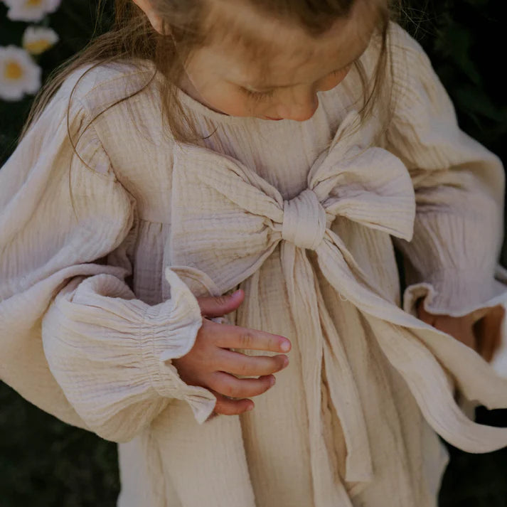 Child wearing a beige dress with a large bow in a natural setting