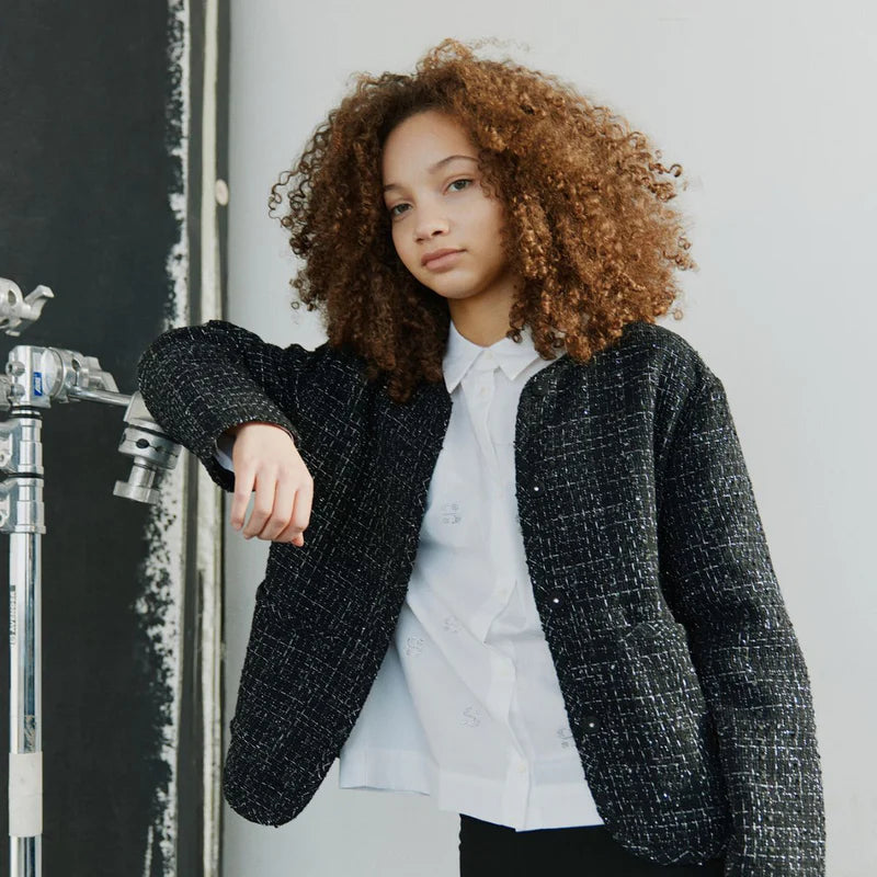 Person wearing a black textured blazer over a white shirt and black pants, standing against a neutral background.